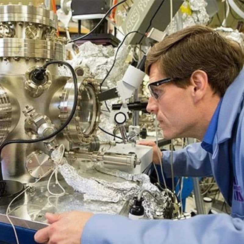 material scientist 800x800 px A material scientist wearing safety glasses examines a complex piece of laboratory equipment with various wires and metallic components, showcasing the exciting work found in chemistry careers.